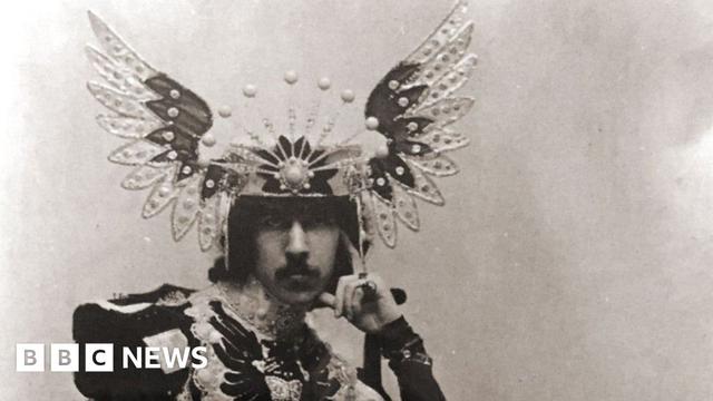 The 5th Marquess of Anglesey in a sepia photograph wearing a floor length overcoat consisting of differently coloured pieces of heavy material. The top of the coat is military in style with large shoulder pieces. He wears a headdress of splayed wings with a tiara-effect frontspiece.