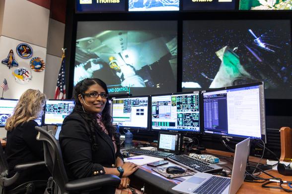 A woman sits at a desk and poses in front of three monitors. Multiple monitors display live data and video of astronauts on the International Space Station. An American flag and NASA mission patches are visible on the wall behind her. A woman sits at a desk and poses in front of three monitors. Multiple monitors display live data and video of astronauts on the International Space Station. An American flag and NASA mission patches are visible on the wall behind her.
