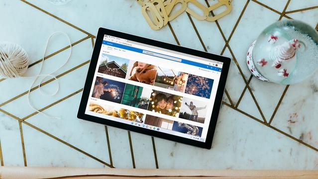 A tablet displaying a photo gallery is placed on a geometric pattern table next to a snow globe, beige yarn, a water bottle, and some gold keychains.