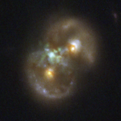 A pair of distant galaxies that form the rough shape of an infinity symbol seen at roughly a 45-degree angle. Two overlapping, fuzzy rings with brighter blue patches are at upper right and lower left. At the center of each ring is a bright yellow blob, which is the nucleus. Where the two rings overlap on the left side, there is a mottled green patch of glowing gas midway between the two yellow nuclei. It is offset slightly to the left.