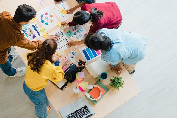 An image of male and female colleagues huddled around a table critiquing each others work. An image of male and female colleagues huddled around a table critiquing each others work.