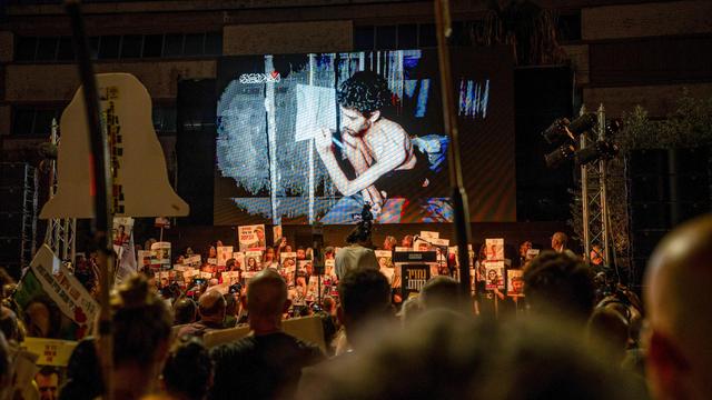 Es ist dunkel. In der unteren Bildhälfte sieht man beleuchtet eine große Menge Demonstranten mit Transparenten und Schildern. Sie schauen auf eine Leinwand über ihnen, auf der das Video zu sehen ist. Man sieht die ausgemergelte Geisel in einem engen Tunnel. 