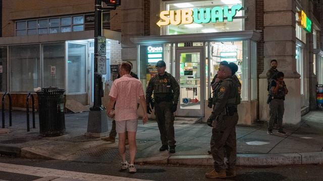 Night scene outside a Subway restaurant with several law enforcement officers in tactical gear and a man in a pink shirt and shorts standing on the sidewalk near a crosswalk.