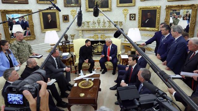 Two men in conversation seated on cream-colored chairs facing each other in an ornate room with gold-framed portraits, surrounded by people, cameras, and microphones overhead.