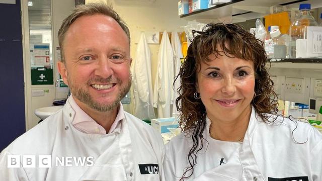 Two scientists, Professors Ed Wild and Sarah Tabrizi, are pictured from the shoulders up. They are both smiling and wearing white lab coats. Prof Wild on the left has a tightly cropped beard. Prof Tabrizi on the right has long wavy hair. They are clearly in a laboratory with scientific equipment on shelves in the background. Two scientists, Professors Ed Wild and Sarah Tabrizi, are pictured from the shoulders up. They are both smiling and wearing white lab coats. Prof Wild on the left has a tightly cropped beard. Prof Tabrizi on the right has long wavy hair. They are clearly in a laboratory with scientific equipment on shelves in the background.