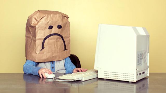 Young Boy in Front of Computer wearing frowny paper bag over head.