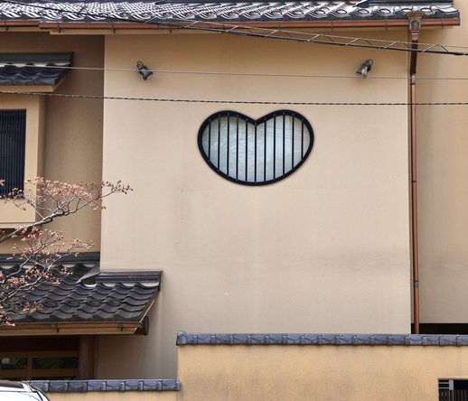 eine hellbraune Hauswand mit einem kleinen Fenster in Herzform, das vergittert ist, jeweils rechts und links oben ist ein Lichtstrahler, links in das Bild ragt einige blühende Kirschblütenzweige hinein, oberhalb des Fensters sind Stromleitungen