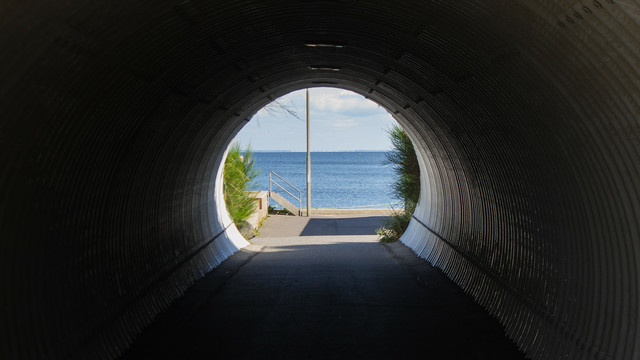 Ein kurzer dunkler Tunnel, an dessen Ende ein Gewässer (Ringkøbing Fjord) bei hellem Sonnenschein zu sehen ist.
