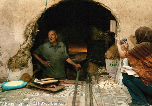 In einer Wand klafft ein Loch, dahinter ein Kellerraum resp. Backstube mit Ofen. Das ganze ist in Marrakesch. Der Bäcker hat soeben 2 frische Brotfladen aus dem Ofen geholt. seine Backschaufel ruht auf dem Ofenrand. Rechts vor dem Loch kniet eine Frau die mit Ihrem Mobiltelefon ein Foto macht