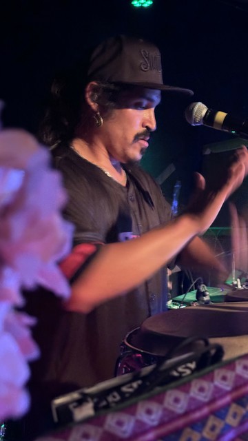 Percussionist wearing a dark baseball cap and shirt, performing on stage with a drum, microphone, and other musical equipment.