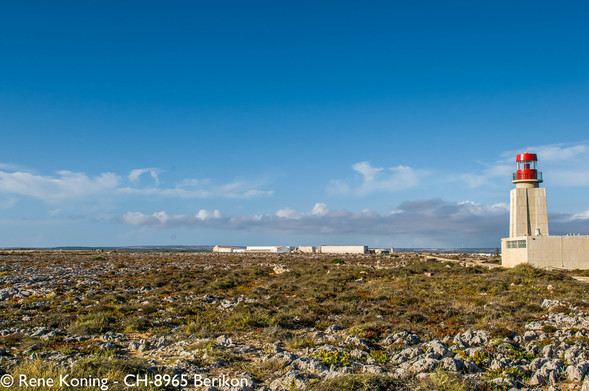 Eine felsige, moosbedeckte,  LAndschaft erstreckt sich zum Horizont. Der Himmel ist Blau und ein paar wenige Cumuluswolken zeigen sich. Am Horizont stehen einstöckige steinbauten. Vom Rechten Bildrand her schaut eine Mauer in das Bild hinein an deren Ende ein Leuchtturm in die höhe ragt. Der Turm ist maximal 10 meter hoch und hat den bekannte roten Aufsatz worin sich das Licht befindet.