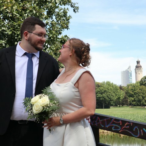 My wife and I on our wedding day, standing on a bridge with a park and Leipzig's distinctive towers in the background. It's a sunny day with blue sky. We're looking each other in the eyes and smiling. She's wearing a modern wedding dress in white satin and is holding a small bouquet. I'm wearing a black suit with a white shirt and blue tie.