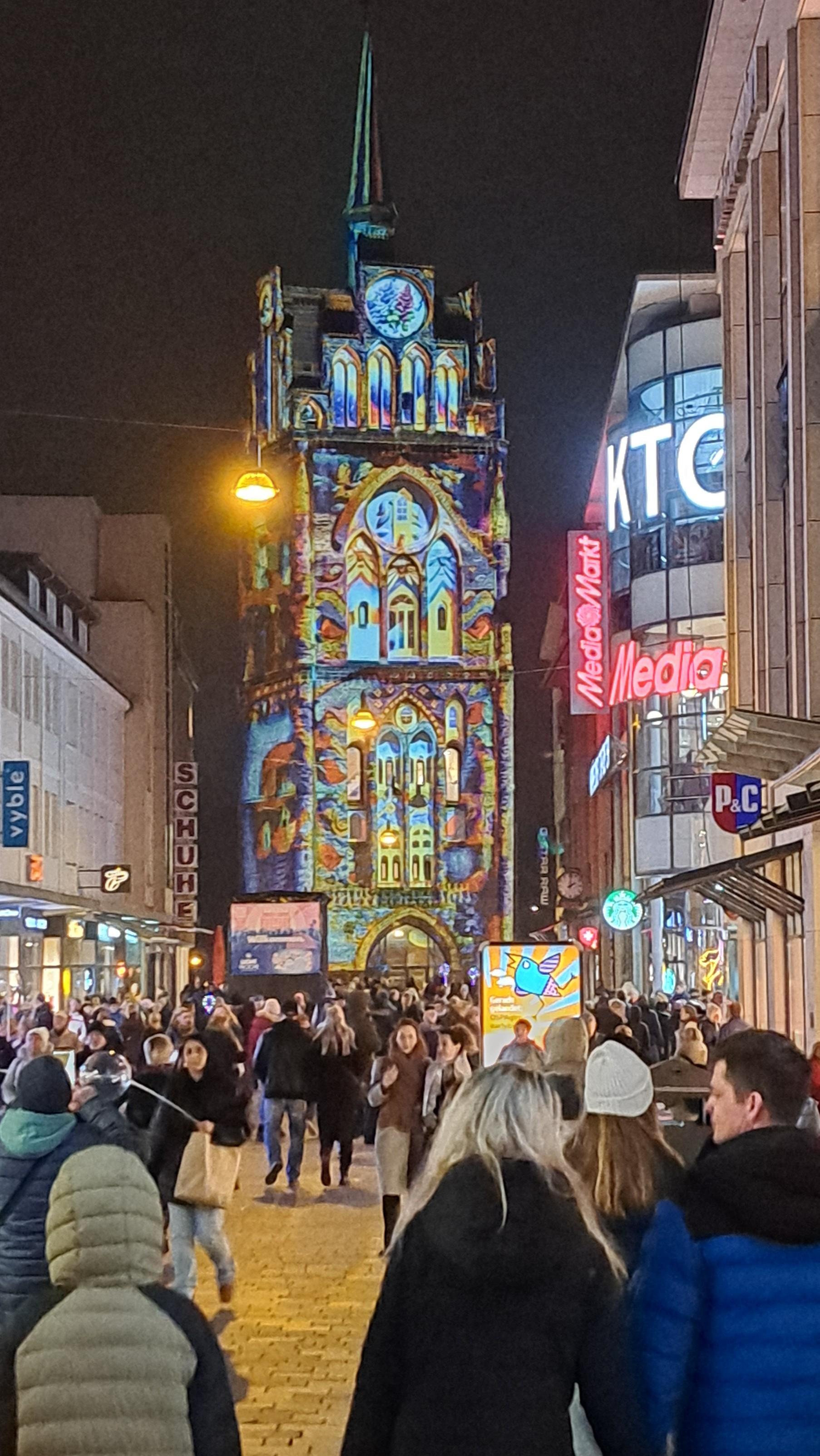 Hochformat, abends einige Stunden nach Sonnenunterganggang. Ein mit bunten Motiven angeleuchtetes hohes mittelalterliches Stadttor. Davor die belebte Fußgängerzone, links und rechts gesäumt von beleuchteten Häusern und Läden.