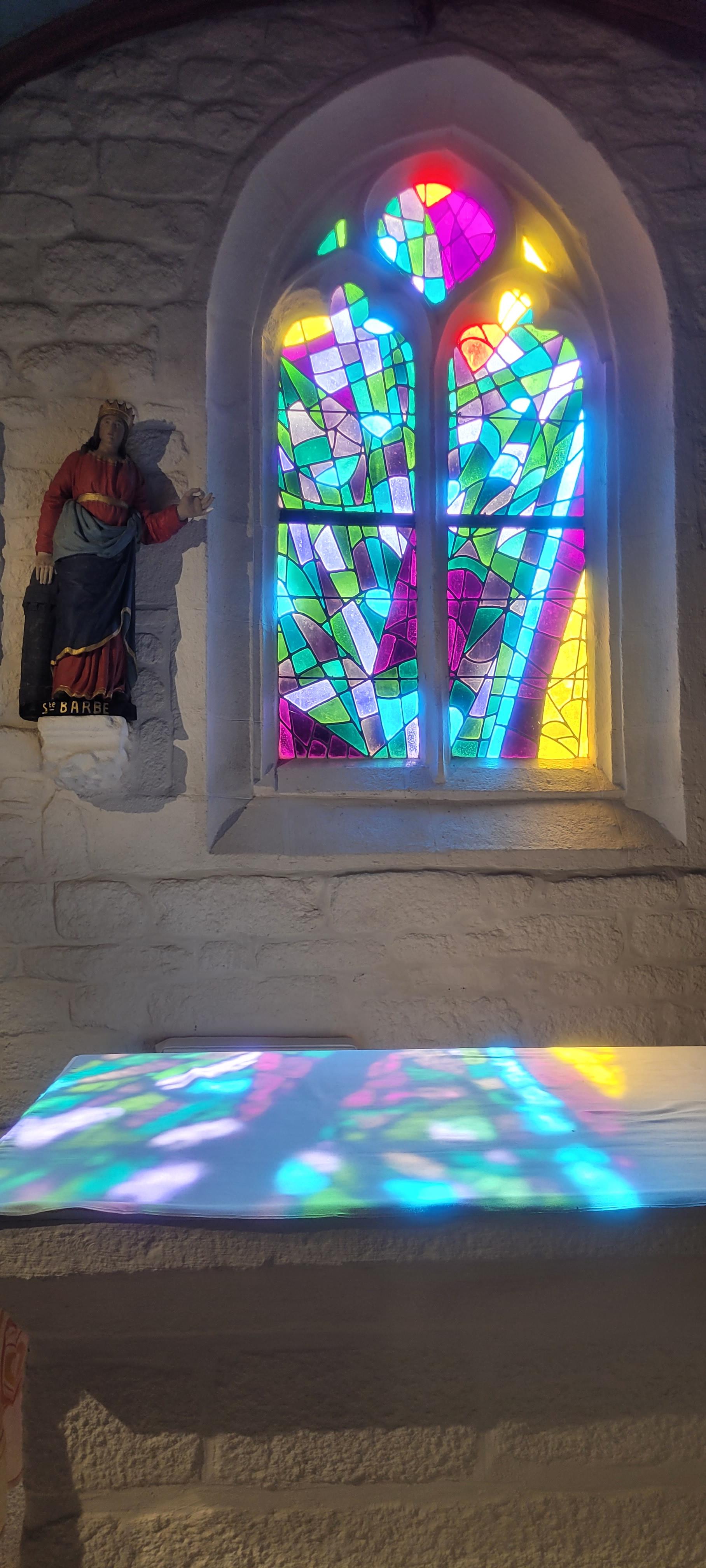 Sonnenlicht fällt duch ein gotisch geformtes Kirchenfenster, in dem Türkis-, Grün-, Violett- und Gelbtöne dominieren und färbt ein weißes Tuch auf dem davor stehenden Altar ein. Daneben eine Statue der Heiligen Barbara.