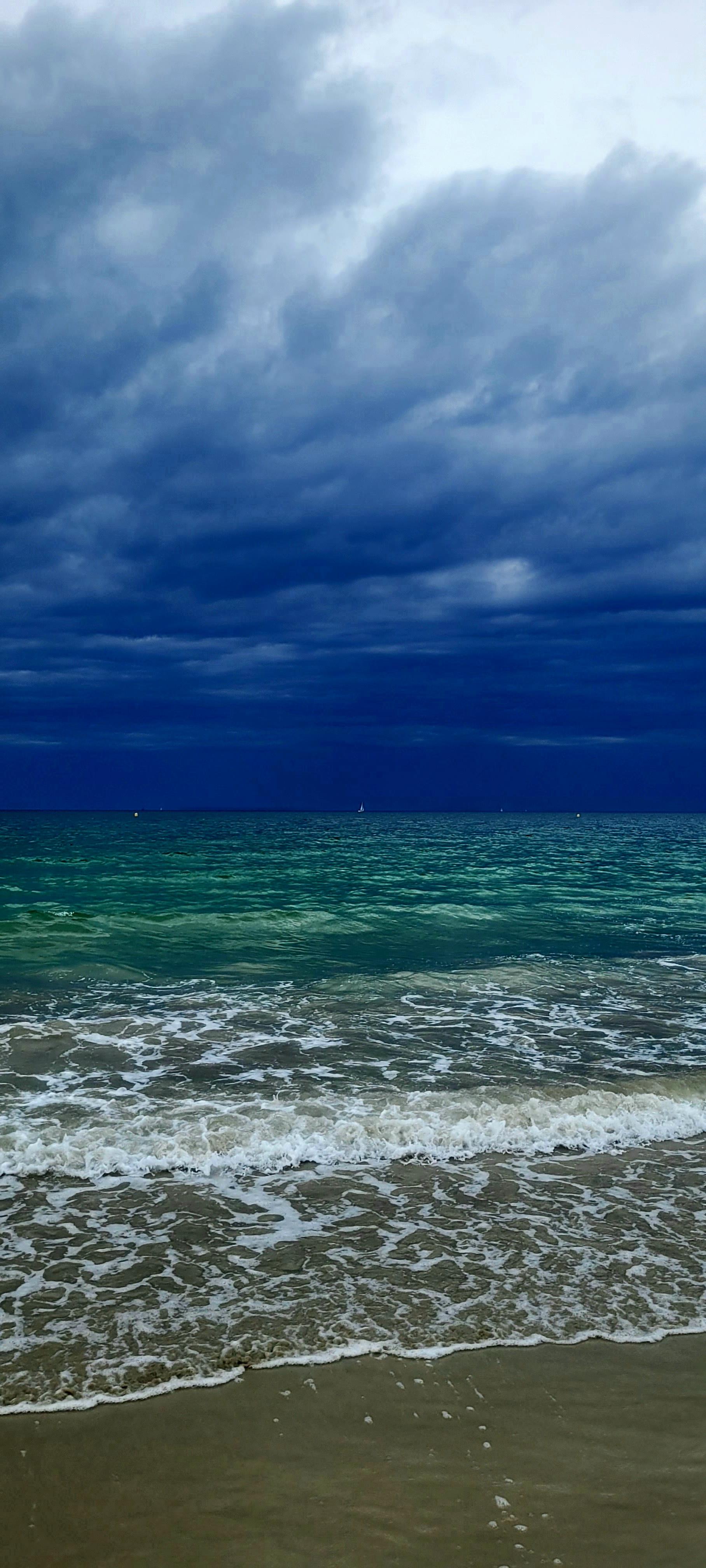 Blick vom Strand auf den Atlantik kurz vor einem Gewitter. Ein dunkelblauen wolkiger Himmel über grünem Wasser, davor Wellenschaum Dr auf den Strand läuft. Am Horizont drei winzige weiße Segel.