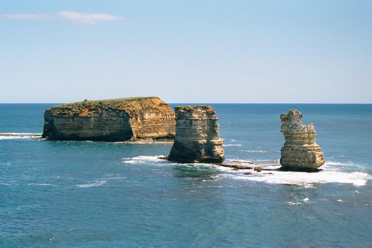 Large rock formations rise from the ocean near the coastline. The largest formation has a flat, grassy top, while the others are more slender and irregular. Gentle waves break around the rocks, and the sky is mostly clear with a few clouds near the horizon.
